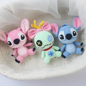 May include: Three handmade crochet dolls in a basket. One is pink, one is mint green, and one is blue. Each doll has large ears, black eyes, and small arms and legs. The pink doll has a pink bow, the green doll has a red collar.