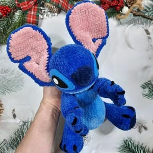 May include: A blue plush toy resembling the character Stitch, with large pink and blue ears, black eyes, and black claws. The toy is made of a soft, textured material and is held by a hand.