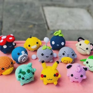May include: A collection of twelve small, round, crocheted plush figures featuring various colors and designs, including a yellow character with red cheeks, a pink character, a blue character with a white spiral, and a red-capped mushroom figure.