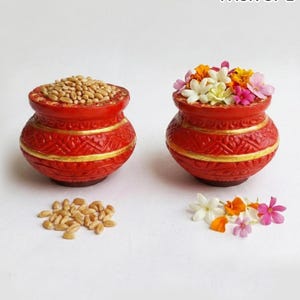 May include: Two round, red clay pots with gold-painted horizontal bands and embossed geometric patterns. One pot is filled with light brown wheat grains, and the other contains a colourful arrangement of white, yellow, and pink flowers.
