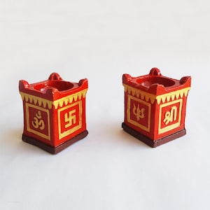 Puede incluir: Dos candelabros cuadrados decorativos rojos y dorados. Cada uno presenta un símbolo religioso en un marco cuadrado dorado. Los bordes superiores son festoneados. La base es marrón.