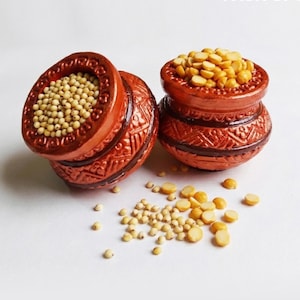 May include: Two small, round, terracotta-colored clay pots with intricate embossed geometric patterns. One pot is filled with small, round, cream-colored grains, and the other is filled with split yellow lentils, with some scattered on the surface.
