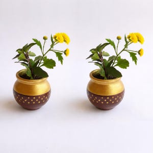 Op de afbeelding: Twee kleine, ronde bloempotten met gele bloemen en groene bladeren. De potten zijn beschilderd met een gouden bovenkant en een bruine onderkant met gouden stippen. De achtergrond is wit.