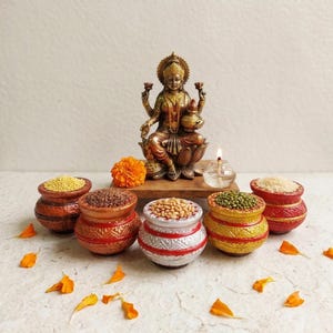 May include: A bronze-toned statue of a deity seated on a lotus base, positioned behind five small, round, decorative pots filled with various grains and seeds. A small glass oil lamp with a lit flame sits next to the statue, with orange marigold petals.