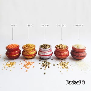 May include: A pack of 5 small, round, decorative pots in red, gold, silver, bronze, and copper finishes. Each pot is filled with different grains or legumes, including lentils and rice, with some scattered on the surface below the pots.