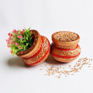 May include: Two round, terracotta-colored pots with embossed geometric patterns and red painted bands. One pot is tilted and filled with small pink artificial flowers and green leaves, while the other stands upright, filled with light brown grains.