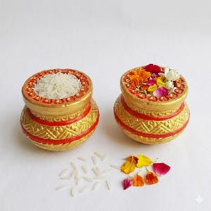 May include: Two small, round, decorative pots painted in metallic gold with bright red horizontal bands and textured geometric patterns. One pot is filled with white rice grains, while the other contains a colourful mix of flower petals.
