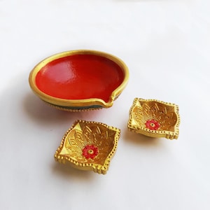 May include: Three decorative oil lamps. One large, round lamp with a red interior and gold trim, and two smaller square lamps with gold exteriors and red floral accents. The lamps are on a white surface.