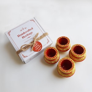 May include: A set of four small, red and gold decorative bowls with ornate detailing. The bowls are arranged near a white box tied with twine, labeled "Handcrafted Heritage."