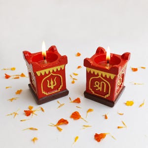 May include: Two square red candle holders with dark brown bases and gold trim. Each holder features a gold embossed symbol on the front and a lit candle inside. The left holder displays a trident, while the right holder shows Sanskrit script.
