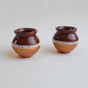 May include: Two small, handcrafted terracotta pots. The upper section is a rich brown, while the lower half is a warm, golden hue. A white band with small, evenly spaced dots encircles each pot, adding a decorative touch.
