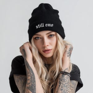 May include: Black beanie with the text "still emo" in white gothic font. The beanie is made of a soft, knitted material. The beanie is worn by a person with blonde hair and tattoos.