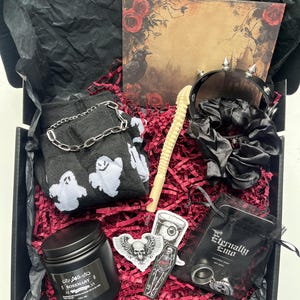 May include: A gift box containing black ghost-patterned socks with a silver chain, a spiked black choker, a black scrunchie, a beige wand, a rosemary candle, skull stickers, and a black pouch labeled Eternally Emo with a skull ring inside.