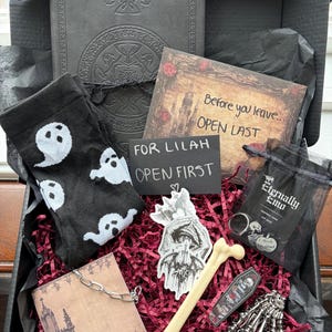 May include: A black gift box containing a black journal with embossed patterns, black socks with white ghost prints, a card reading Before you leave... OPEN LAST, a note reading FOR LILAH OPEN FIRST, a bone-shaped pen, a skeleton hand bracelet, and a small bag.