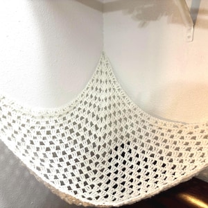 May include: White crocheted hammock hanging in a corner of a room. The hammock is made of a delicate, open weave and is perfect for a small pet or child.