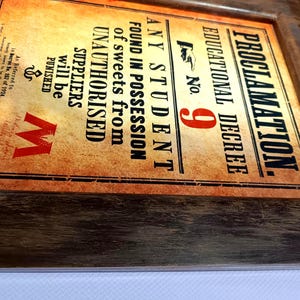 Può includere: Stampa incorniciata dall'aspetto di pergamena vintage. Il testo recita "PROCLAMAZIONE. DECRETO EDUCATIVO n. 9" e mette in guardia dai dolci non autorizzati. La cornice è in legno scuro e invecchiato. È visibile un logo "M" rosso.