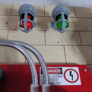 May include: Two gray electrical warning lights with red and green bulbs mounted on a wall. A red electrical panel with a black and white warning sign that reads "DANGER ELECTRICAL EQUIPMENT AUTHORIZED PERSONNEL ONLY" is below the lights. Two silver flexible conduit pipes are connected to the panel.
