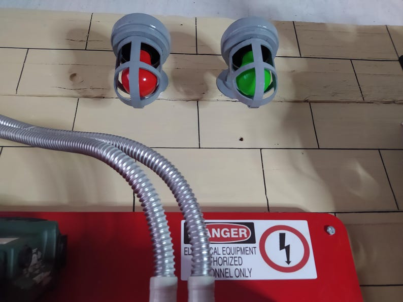 May include: Two gray electrical warning lights with red and green bulbs mounted on a wall. A red electrical panel with a black and white warning sign that reads "DANGER ELECTRICAL EQUIPMENT AUTHORIZED PERSONNEL ONLY" is below the lights. Two silver flexible conduit pipes are connected to the panel.