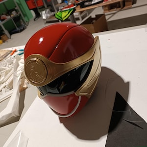 Könnte beinhalten: Ein roter und goldener Power Ranger Helm mit schwarzem Visier. Der Helm hat einen goldenen Rand und ein goldenes Emblem auf der Vorderseite.