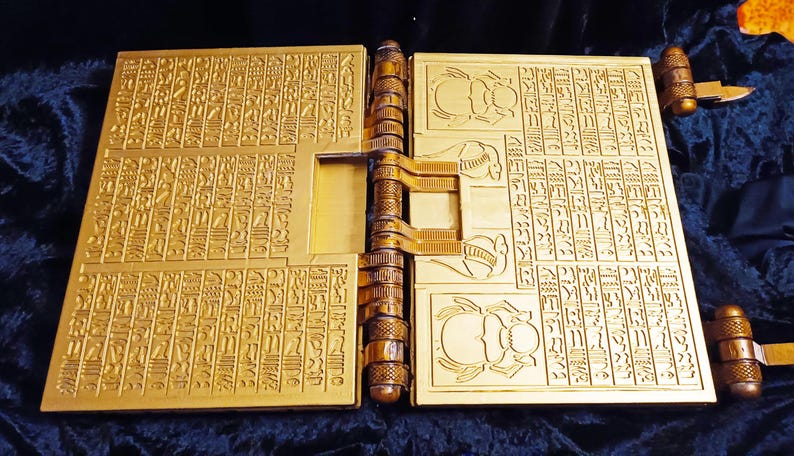 Puede incluir: Un libro de color dorado con un dise&ntilde;o detallado e intrincado en la portada. El libro est&aacute; cerrado y tiene una bisagra con un dise&ntilde;o decorativo. El libro est&aacute; sobre una superficie azul oscuro.
