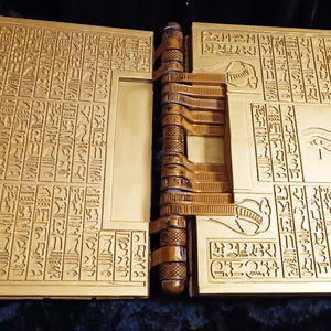 Puede incluir: Un libro de color dorado con un dise&ntilde;o detallado e intrincado de jerogl&iacute;ficos y un s&iacute;mbolo del ojo de Horus. El libro est&aacute; cerrado y tiene una bisagra decorativa.