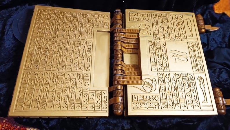 Puede incluir: Un libro de color dorado con un dise&ntilde;o detallado e intrincado de jerogl&iacute;ficos y un s&iacute;mbolo del ojo de Horus. El libro est&aacute; cerrado y tiene una bisagra decorativa.