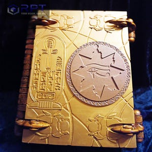 Puede incluir: Un libro de color dorado con un dise&ntilde;o intrincado que presenta un s&iacute;mbolo ankh egipcio, una estrella con un ojo y escarabajos. El libro tiene un lomo con una serie de protuberancias elevadas.