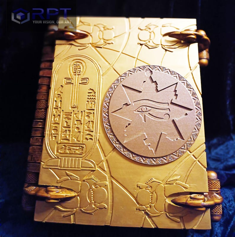 Puede incluir: Un libro de color dorado con un dise&ntilde;o intrincado que presenta un s&iacute;mbolo ankh egipcio, una estrella con un ojo y escarabajos. El libro tiene un lomo con una serie de protuberancias elevadas.