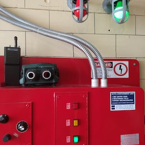 May include: A red metal control panel with a green, yellow, and red light. The panel has a sticker that reads "ECTO-CONTAINMENT UNIT 1: SET ENTRY GRID 2: NEUTRINIZE FIELD 3: GHOST INCARCERATED" and a warning about loss of power. There are two gray light fixtures with red and green lights on the wall above the panel.