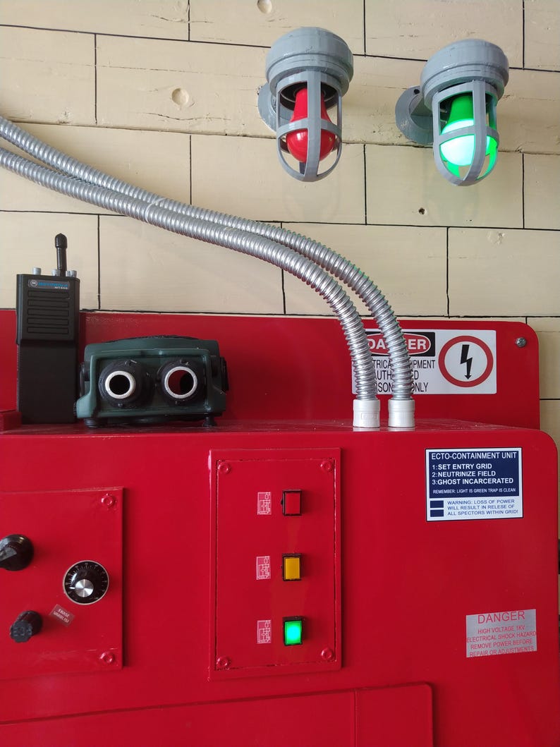 May include: A red metal control panel with a green, yellow, and red light. The panel has a sticker that reads "ECTO-CONTAINMENT UNIT 1: SET ENTRY GRID 2: NEUTRINIZE FIELD 3: GHOST INCARCERATED" and a warning about loss of power. There are two gray light fixtures with red and green lights on the wall above the panel.