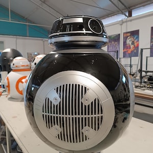 May include: A large, black and silver BB-series droid prop with a spherical body and a domed head. The droid features a silver grill on the front and is displayed in a workshop setting. Other droids are visible in the background.