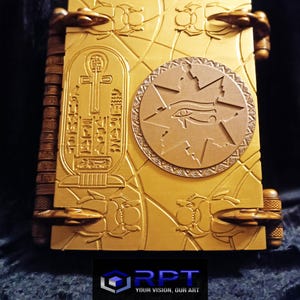 Puede incluir: Un libro de color dorado con intrincados tallados de escarabajos, un ankh y un ojo de Horus. El libro tiene un lomo de cuero y cierres met&aacute;licos. El texto en la portada dice "RPT YOUR VISION, OUR ART".
