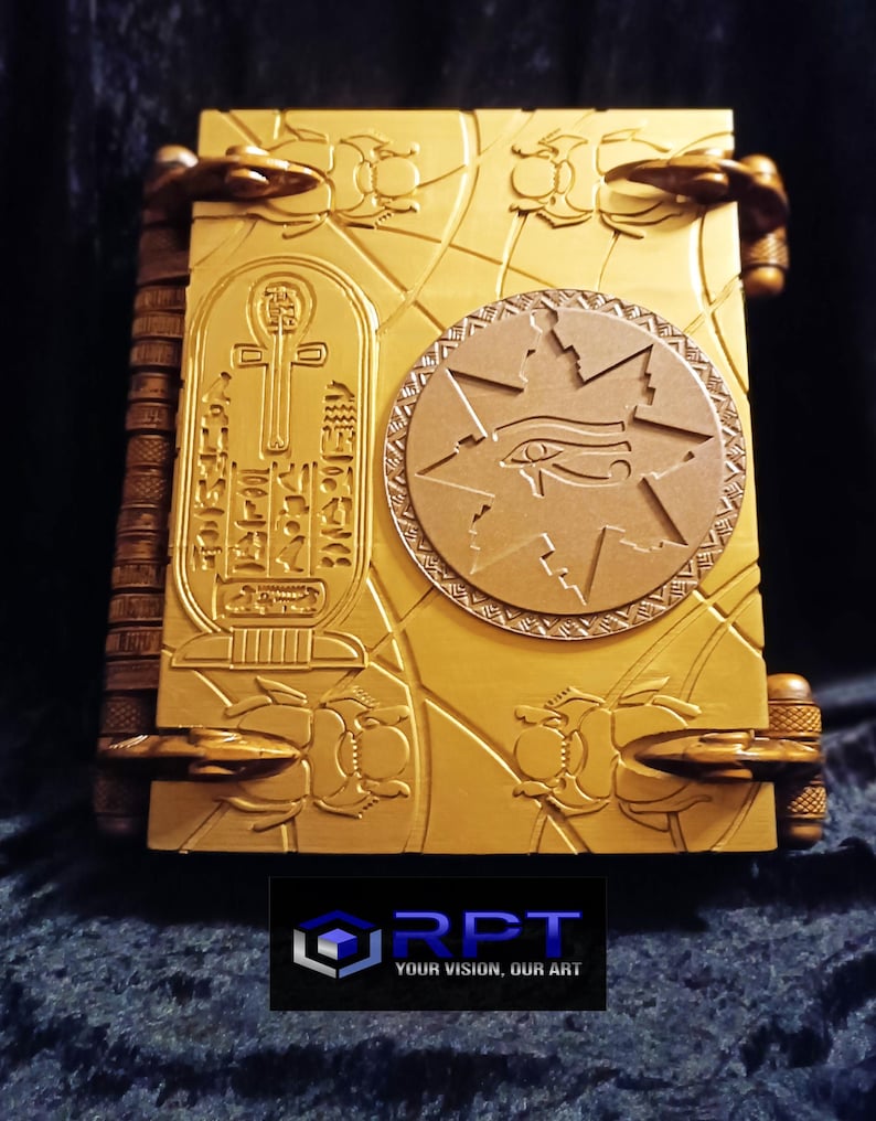 Puede incluir: Un libro de color dorado con intrincados tallados de escarabajos, un ankh y un ojo de Horus. El libro tiene un lomo de cuero y cierres met&aacute;licos. El texto en la portada dice "RPT YOUR VISION, OUR ART".