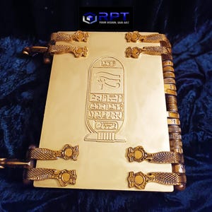 Puede incluir: Un libro de color dorado con un dise&ntilde;o de estilo egipcio. La portada presenta un s&iacute;mbolo jerogl&iacute;fico y un ojo alado. El libro tiene un lomo decorativo con una serie de anillos met&aacute;licos.