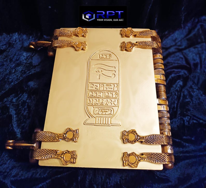 Puede incluir: Un libro de color dorado con un dise&ntilde;o de estilo egipcio. La portada presenta un s&iacute;mbolo jerogl&iacute;fico y un ojo alado. El libro tiene un lomo decorativo con una serie de anillos met&aacute;licos.