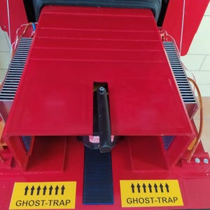 May include: A red metal contraption with two black cooling fins on either side. The contraption has a black handle and a black platform in the center. The platform has a blue and black striped surface. The contraption has two yellow labels on the front that say "GHOST-TRAP" with arrows pointing upwards.