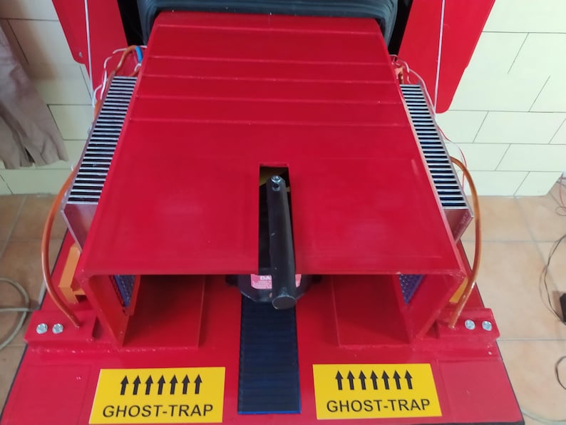May include: A red metal contraption with two black cooling fins on either side. The contraption has a black handle and a black platform in the center. The platform has a blue and black striped surface. The contraption has two yellow labels on the front that say "GHOST-TRAP" with arrows pointing upwards.