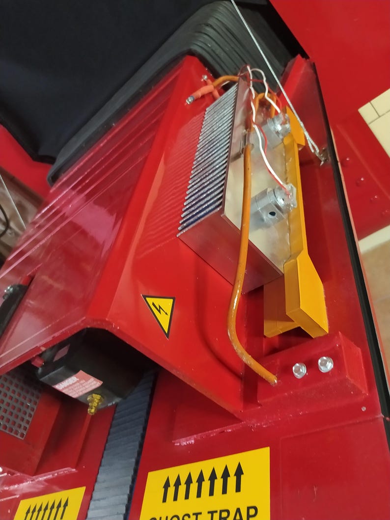 May include: A red metal machine with a yellow warning sign and a black and white warning sign that says "Ghost Trap". The machine has a silver heat sink with wires attached to it.
