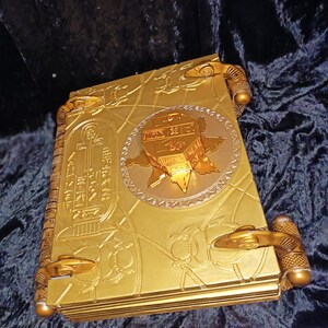 Puede incluir: Un libro de color dorado con dise&ntilde;os intrincados y un emblema circular dorado ornamentado en relieve en la portada. El libro tiene cierres decorativos dorados en los lados.
