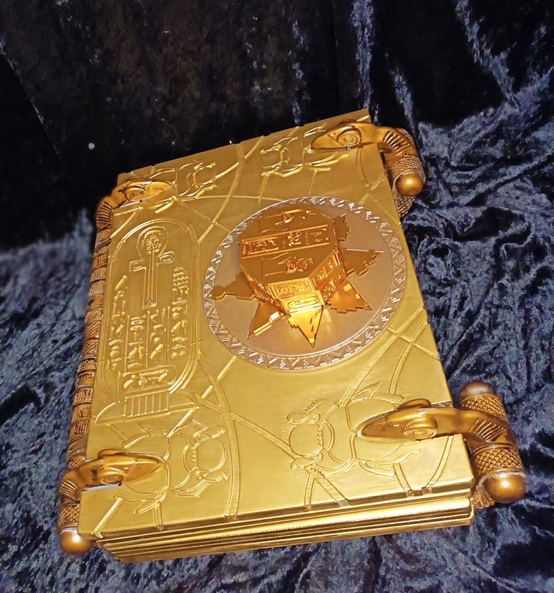 Puede incluir: Un libro de color dorado con dise&ntilde;os intrincados y un emblema circular dorado ornamentado en relieve en la portada. El libro tiene cierres decorativos dorados en los lados.