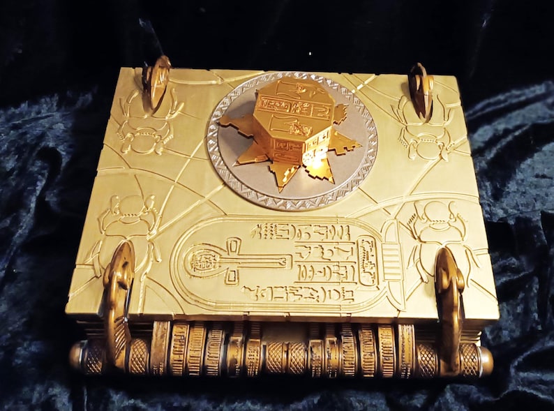 Puede incluir: Un libro de color dorado con un dise&ntilde;o decorativo e intrincado. El libro tiene una parte superior en forma de estrella de color dorado con jerogl&iacute;ficos. El libro tiene dos asas doradas en los lados. El libro est&aacute; cerrado y tiene una serie de anillos dorados en el lomo.