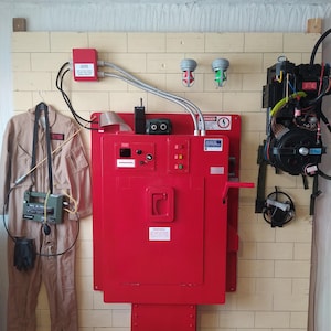 Puede incluir: Una caja de metal roja con una puerta y una manija. La caja tiene una señal de advertencia que dice "PELIGRO ALTA TENSIÓN". Hay dos luces verdes y una luz roja en la caja. Hay un mono de trabajo y un dispositivo de caza de fantasmas colgando en la pared al lado de la caja.