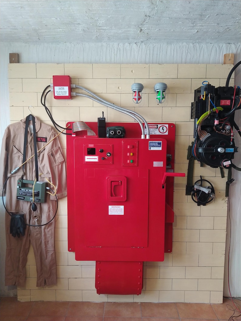 May include: A red metal box with a door and a handle. The box has a warning sign that says "DANGER HIGH VOLTAGE". There are two green lights and one red light on the box. There is a jumpsuit and a ghost hunting device hanging on the wall next to the box.