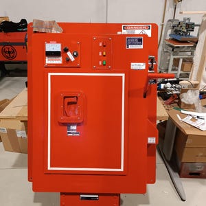 Puede incluir: Un gran panel eléctrico rojo con un borde blanco y varios medidores, interruptores y etiquetas de advertencia. El panel tiene una manija y una sección inferior. La palabra "DANGER" es visible en una etiqueta. El panel está en un taller.