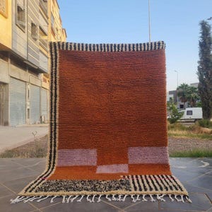 Puede incluir: Una alfombra rectangular, texturizada, naranja con bordes y flecos a rayas blancas y negras. La alfombra presenta formas geométricas en tonos morados y naranjas. La alfombra se exhibe al aire libre contra un edificio.