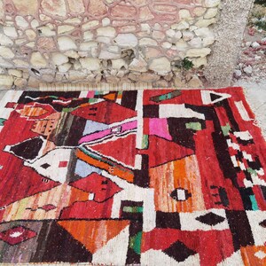 May include: A colorful, abstract rug with geometric patterns in shades of red, orange, black, white, and green. The rug is woven with a variety of textures and materials, creating a unique and eye-catching design.
