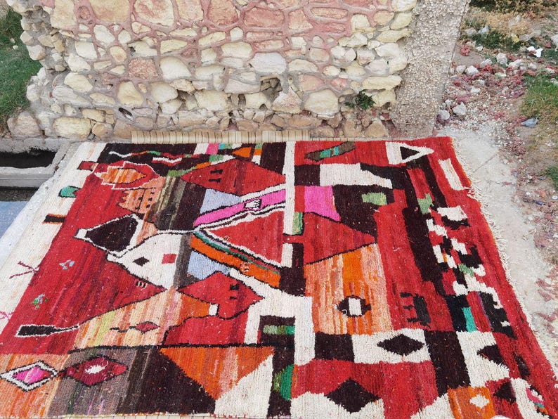 May include: A colorful, abstract rug with geometric patterns in shades of red, orange, black, white, and green. The rug is woven with a variety of textures and materials, creating a unique and eye-catching design.