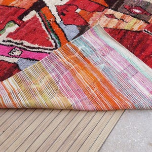May include: A colorful Moroccan rug with a geometric pattern in shades of red, pink, orange, and white. The rug is folded in half, revealing the woven backing.