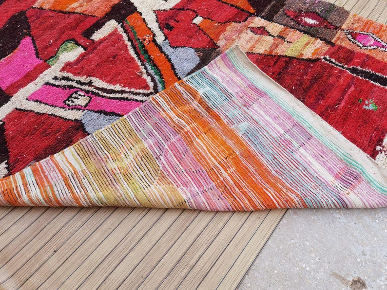 May include: A colorful Moroccan rug with a geometric pattern in shades of red, pink, orange, and white. The rug is folded in half, revealing the woven backing.