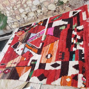 May include: A colorful Moroccan rug with geometric patterns in shades of red, orange, white, and black. The rug has a fringed edge and is made of wool.